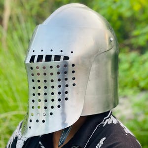K&ouml;nnte beinhalten: Ein polierter silberner Ritterhelm mit einem Visier mit zahlreichen kleinen, gleichm&auml;&szlig;ig verteilten L&ouml;chern. Der Helm hat eine abgerundete Oberseite und einen sch&uuml;tzenden Nackenschutz. Der Hintergrund ist eine verschwommene Ansicht von gr&uuml;nem Laub.