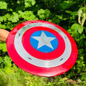 May include: A circular shield in red, white, and blue, featuring a central blue star. The shield has a red outer ring, a silver middle ring, and a blue centre with a white star. The shield is held against a backdrop of green foliage.