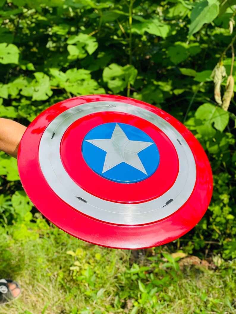 May include: A circular shield in red, white, and blue, featuring a central blue star. The shield has a red outer ring, a silver middle ring, and a blue centre with a white star. The shield is held against a backdrop of green foliage.