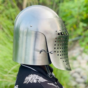 K&ouml;nnte beinhalten: Ein silberfarbener mittelalterlicher Helm mit Visier, das zahlreiche kleine L&ouml;cher zur Bel&uuml;ftung aufweist. Der Helm hat eine abgerundete Oberseite und ein aufklappbares Visier. Der Hintergrund ist ein verschwommenes Gr&uuml;n.