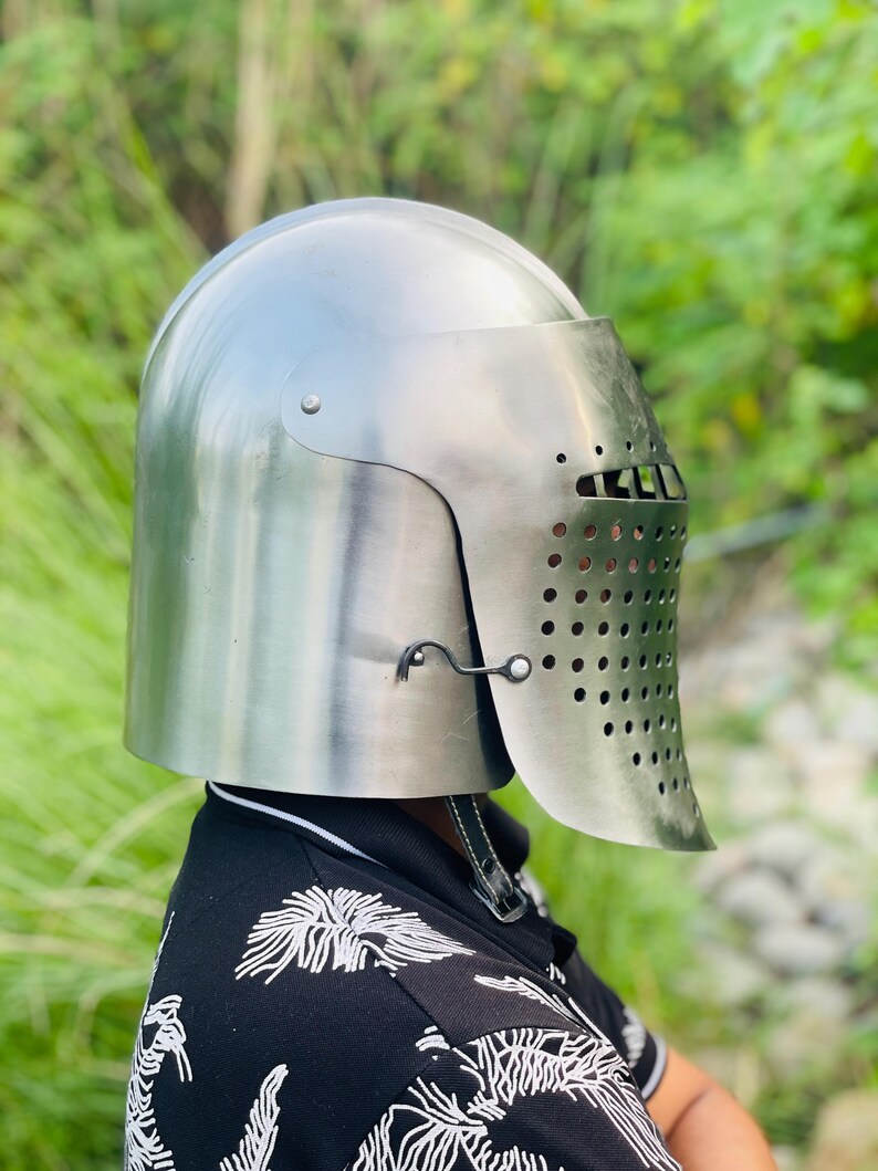 K&ouml;nnte beinhalten: Ein silberfarbener mittelalterlicher Helm mit Visier, das zahlreiche kleine L&ouml;cher zur Bel&uuml;ftung aufweist. Der Helm hat eine abgerundete Oberseite und ein aufklappbares Visier. Der Hintergrund ist ein verschwommenes Gr&uuml;n.