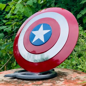 May include: A Captain America shield prop, with a red, white, and blue colour scheme. The shield has a circular design with a white star on a blue centre, surrounded by concentric rings of white and red. The prop is displayed on a black stand.