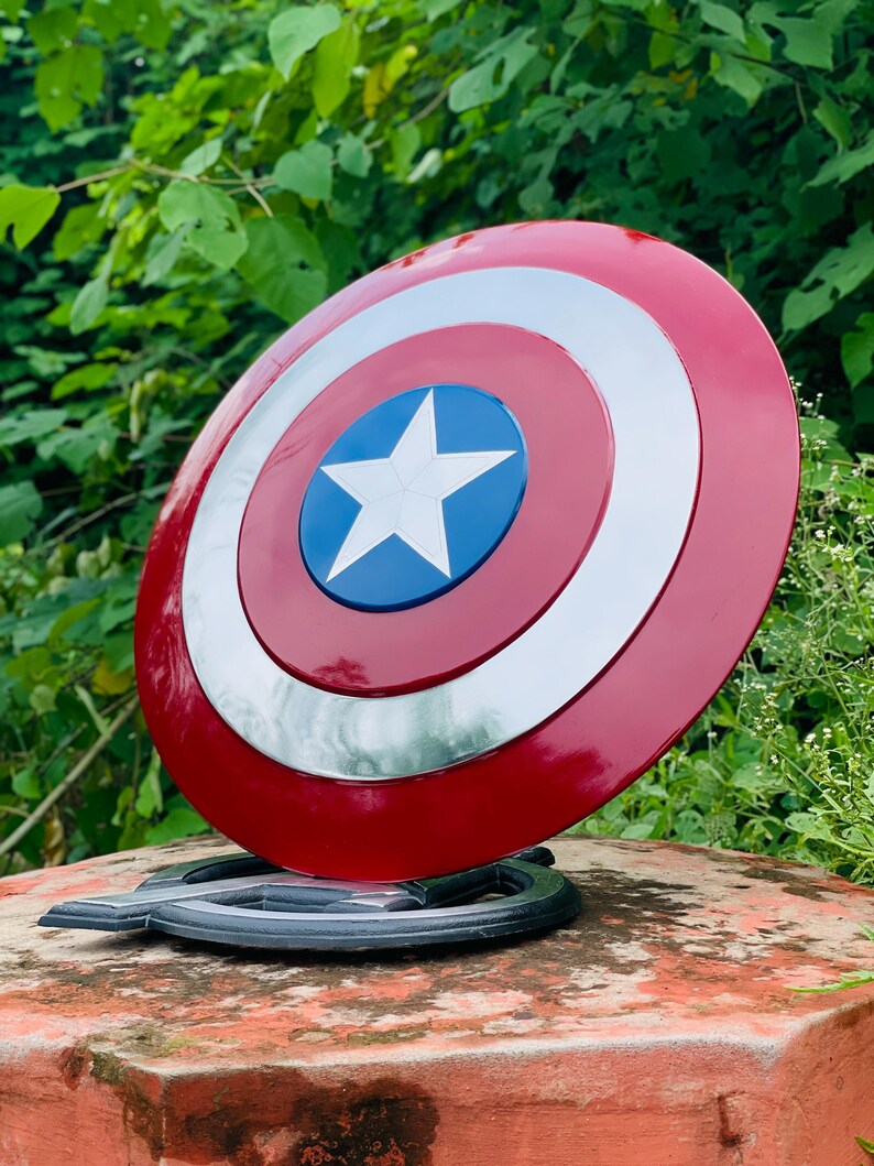 May include: A Captain America shield prop, with a red, white, and blue colour scheme. The shield has a circular design with a white star on a blue centre, surrounded by concentric rings of white and red. The prop is displayed on a black stand.