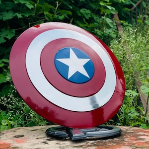 May include: A Captain America shield, with a red, white, and blue design. The shield has a central blue circle with a white star, encircled by white and silver rings. It is mounted on a stand, showcasing its iconic design.
