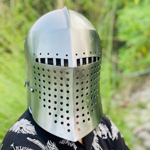 K&ouml;nnte beinhalten: Ein silberfarbener mittelalterlicher Helm mit Visier, das kleine, gleichm&auml;&szlig;ig verteilte L&ouml;cher f&uuml;r Sicht und Bel&uuml;ftung aufweist. Der Helm hat eine abgerundete Oberseite und ein klappbares Visier, das das Gesicht sch&uuml;tzt. Der Hintergrund ist verschwommen, mit gr&uuml;nem Laub.