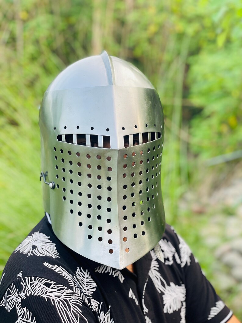 K&ouml;nnte beinhalten: Ein silberfarbener mittelalterlicher Helm mit Visier, das kleine, gleichm&auml;&szlig;ig verteilte L&ouml;cher f&uuml;r Sicht und Bel&uuml;ftung aufweist. Der Helm hat eine abgerundete Oberseite und ein klappbares Visier, das das Gesicht sch&uuml;tzt. Der Hintergrund ist verschwommen, mit gr&uuml;nem Laub.