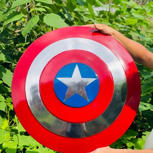 May include: A round shield in red, white, and blue, featuring a silver star and concentric circles. The shield is made of metal. The shield is held up against a backdrop of green foliage.