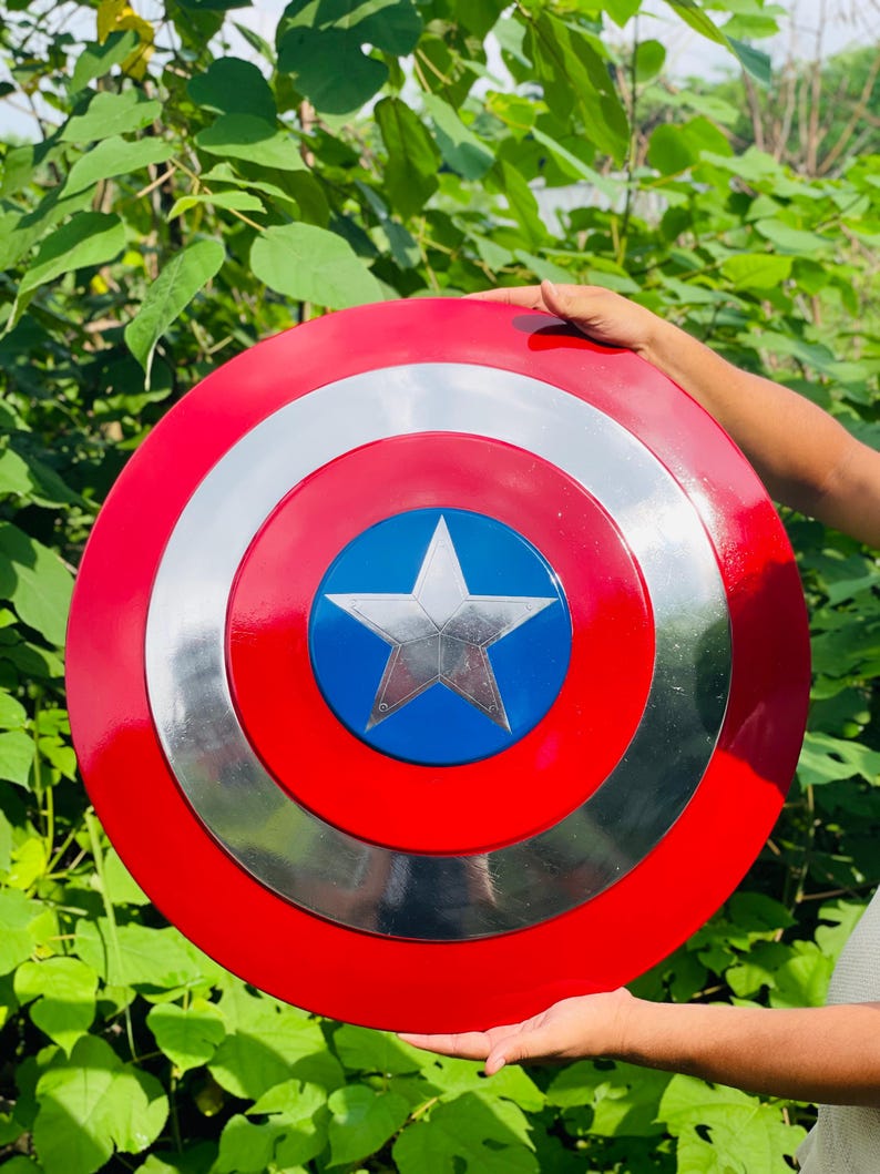 May include: A round shield in red, white, and blue, featuring a silver star and concentric circles. The shield is made of metal. The shield is held up against a backdrop of green foliage.