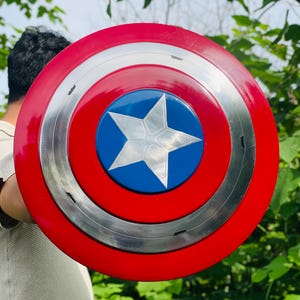 May include: A Captain America shield in red, white, and blue. The shield features a red outer ring, a silver middle ring, and a blue centre with a white star. The round shield appears to be made of metal.
