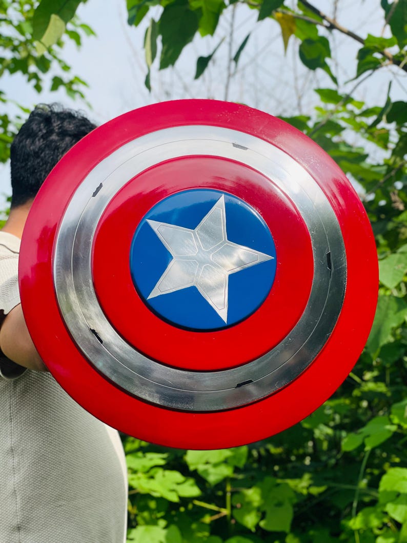 May include: A Captain America shield in red, white, and blue. The shield features a red outer ring, a silver middle ring, and a blue centre with a white star. The round shield appears to be made of metal.