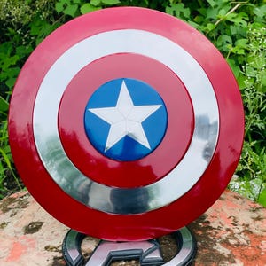 May include: A Captain America shield replica. The shield is circular with a red outer ring, a silver inner ring, and a blue center with a white star. It is displayed on a silver and black Avengers logo base.