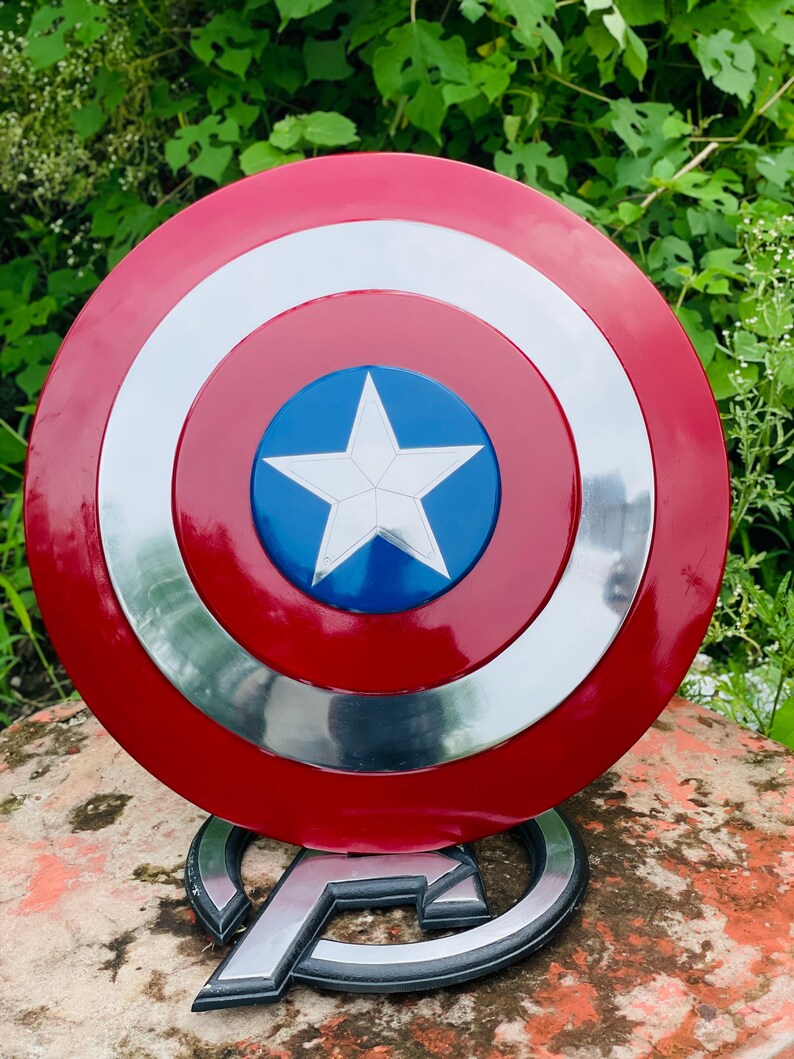May include: A Captain America shield replica. The shield is circular with a red outer ring, a silver inner ring, and a blue center with a white star. It is displayed on a silver and black Avengers logo base.