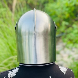 K&ouml;nnte beinhalten: Ein polierter, silberfarbener mittelalterlicher Helm mit abgerundeter Oberseite und einer vertikalen Naht in der Mitte. Der Helm wird &uuml;ber einem schwarzen Hemd mit wei&szlig;em Blumenmuster getragen. Der Hintergrund ist verschwommenes gr&uuml;nes Laub.