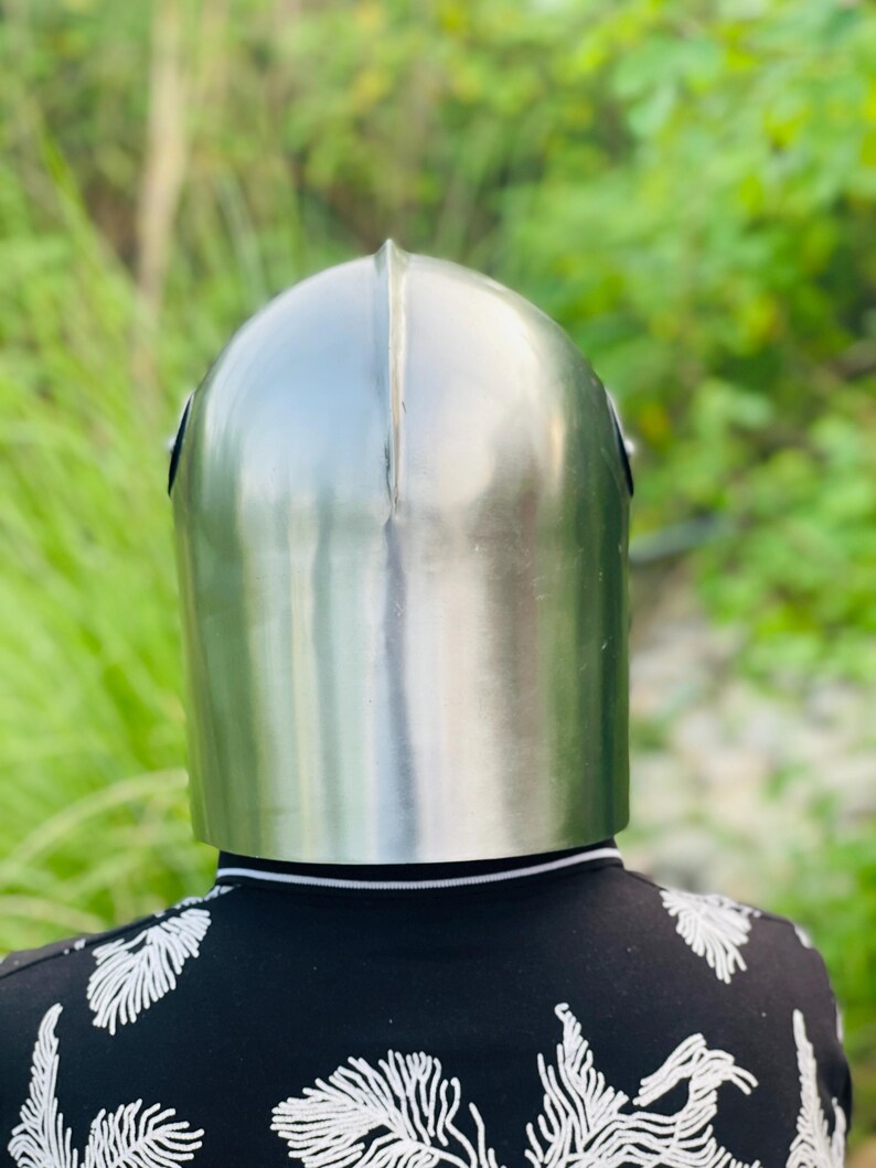 K&ouml;nnte beinhalten: Ein polierter, silberfarbener mittelalterlicher Helm mit abgerundeter Oberseite und einer vertikalen Naht in der Mitte. Der Helm wird &uuml;ber einem schwarzen Hemd mit wei&szlig;em Blumenmuster getragen. Der Hintergrund ist verschwommenes gr&uuml;nes Laub.