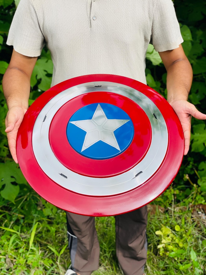May include: A circular shield in red, white, and blue, featuring a silver star at its centre. The shield has a red outer ring, a silver middle ring, and a blue central area with a white star. A classic superhero accessory.