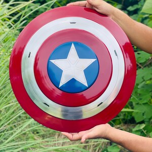 May include: A Captain America shield in red, white, blue, and silver. The shield features a red outer ring, a silver middle ring, and a blue centre with a white star. The round shield appears to be made of metal.
