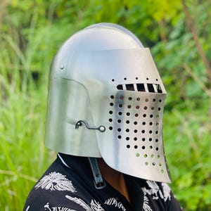 K&ouml;nnte beinhalten: Ein polierter silberner Ritterhelm mit Visier und perforiertem Gesichtsschutz. Der Helm hat eine abgerundete Oberseite und wird mit einem Kinnriemen befestigt. Der Hintergrund ist verschwommenes gr&uuml;nes Laub.