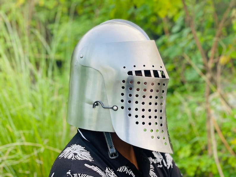 K&ouml;nnte beinhalten: Ein polierter silberner Ritterhelm mit Visier und perforiertem Gesichtsschutz. Der Helm hat eine abgerundete Oberseite und wird mit einem Kinnriemen befestigt. Der Hintergrund ist verschwommenes gr&uuml;nes Laub.