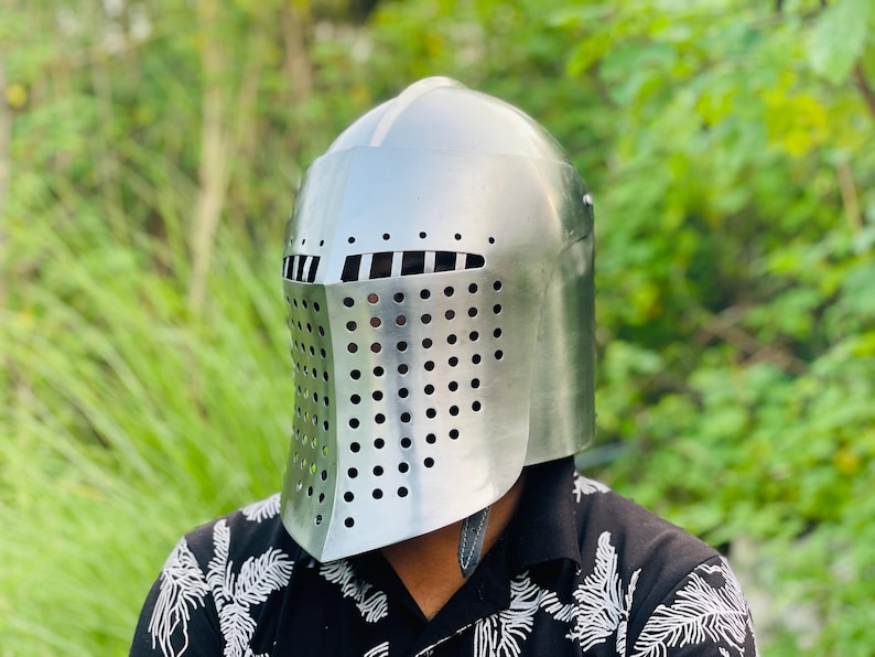 K&ouml;nnte beinhalten: Ein silberfarbener Ritterhelm mit abgerundeter Oberseite und einem Gesichtsschutz mit zahlreichen kleinen, gleichm&auml;&szlig;ig verteilten L&ouml;chern. Der Helm hat ein Visier mit horizontalen Schlitzen. Der Hintergrund ist verschwommenes gr&uuml;nes Laub.
