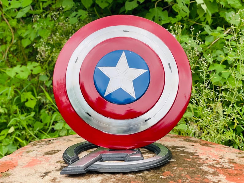 May include: A red, white, and blue Captain America shield with a white star in the centre. The shield is round with silver accents and sits on a black and silver Avengers logo base. The shield is a replica of the one used by the superhero.