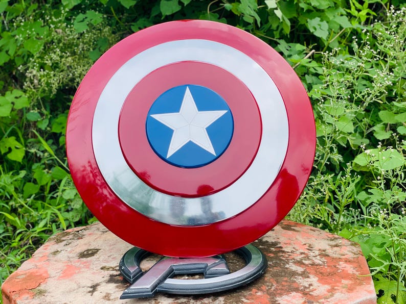 May include: A circular Captain America shield in red, white, and blue, featuring a white star. The shield has concentric rings of red, silver, and white. It is displayed on a silver Avengers logo base.