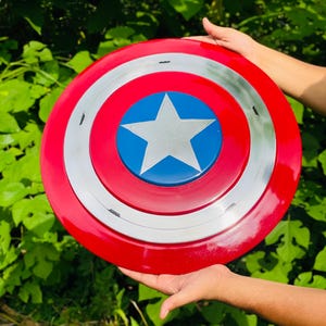 May include: A circular shield in red, white, and blue, featuring a central blue circle with a white star. The design includes a silver band separating the red and blue areas. A classic superhero accessory.