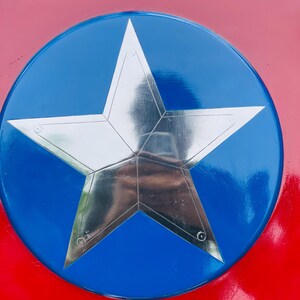 May include: A shield in red, white, and blue, featuring a large, silver five-pointed star at its centre. The star is set against a blue circle, with a red border. The shield has a glossy finish.