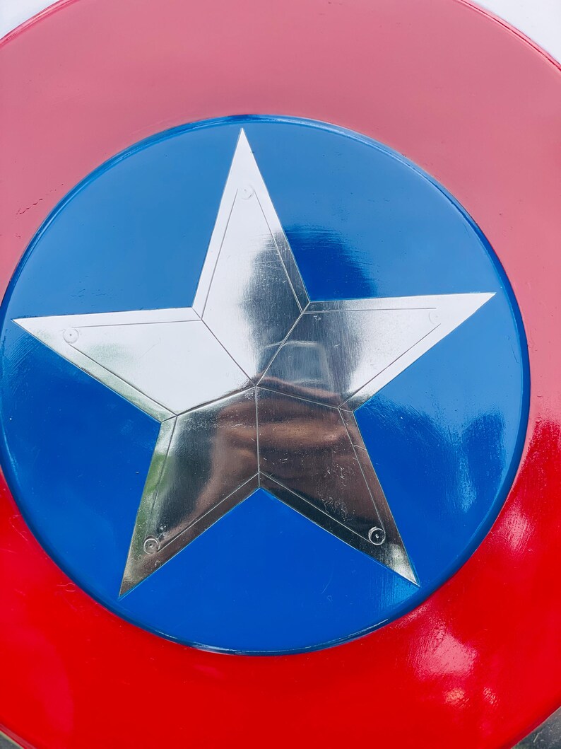 May include: A shield in red, white, and blue, featuring a large, silver five-pointed star at its centre. The star is set against a blue circle, with a red border. The shield has a glossy finish.