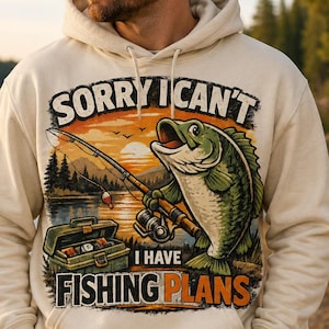 May include: A cream-colored hooded sweatshirt featuring a graphic print of a green fish holding a fishing rod, a tackle box, and the text SORRY I CAN'T I HAVE FISHING PLANS. The design includes a sunset over a lake with trees in the background.