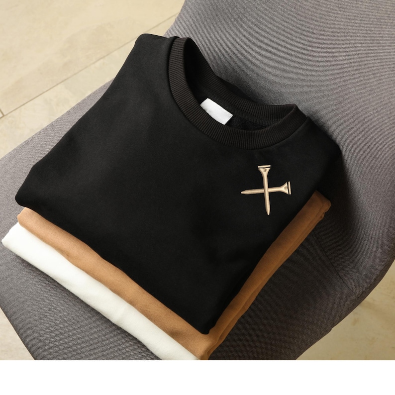Puede incluir: Una pila de tres sudaderas plegadas en negro, beige y blanco. La sudadera negra superior tiene un emblema dorado de tee de golf en el pecho. Las sudaderas est&aacute;n cuidadosamente dispuestas sobre una silla gris.