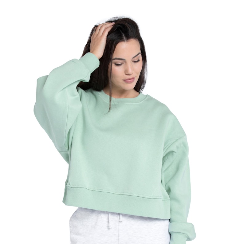 Puede incluir: Una sudadera corta de color verde claro con mangas largas. La sudadera tiene cuello redondo y dobladillo sin rematar. La modelo lleva la sudadera con pantalones de ch&aacute;ndal grises. El fondo es blanco.