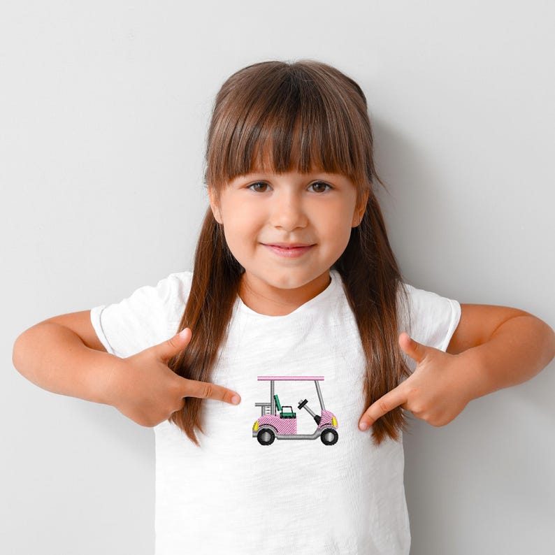 Puede incluir: Una camiseta blanca con un dise&ntilde;o de carrito de golf rosa y amarillo. El carrito de golf tiene un techo rosa, ruedas amarillas y un asiento verde. Una ni&ntilde;a se&ntilde;ala la camiseta.