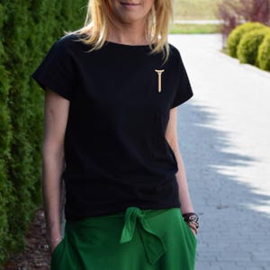Puede incluir: Una camiseta negra con un peque&ntilde;o alfiler dorado en forma de 'T' en el bolsillo del pecho. La persona lleva pantalones verdes. El fondo presenta un seto verde y una pasarela pavimentada.