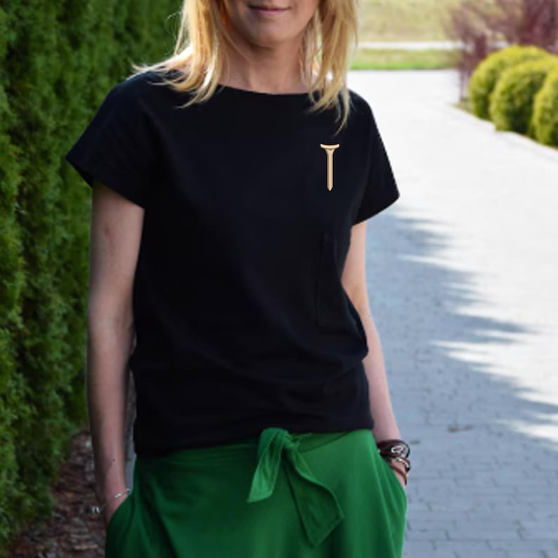 Puede incluir: Una camiseta negra con un peque&ntilde;o alfiler dorado en forma de 'T' en el bolsillo del pecho. La persona lleva pantalones verdes. El fondo presenta un seto verde y una pasarela pavimentada.