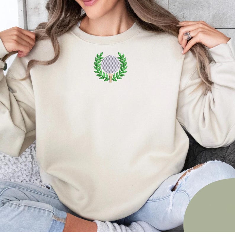 Puede incluir: Sudadera color crema con un dise&ntilde;o de pelota de golf bordada. La pelota de golf est&aacute; rodeada por una corona de laurel verde. La sudadera tiene mangas largas y cuello redondo.