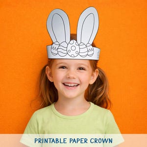 May include: A smiling child wearing a white paper Easter bunny crown with decorative eggs. The crown is printed with black ink and has large bunny ears. The background is a solid orange color. The text "PRINTABLE PAPER CROWN" is at the bottom.