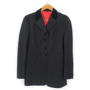 Peut inclure: Un blazer noir avec un col en velours et une doublure rouge, suspendu à un cintre en bois. Le blazer a une fermeture à trois boutons et une poche unique. Le vêtement est présenté sur fond blanc.