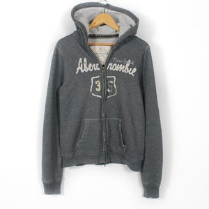 May include: A charcoal grey zip-up hoodie with a drawstring hood and front pockets. The hoodie features the text "Abercrombie New York" in a cursive font, along with a "35" emblem. The jacket is hanging on a wooden hanger.