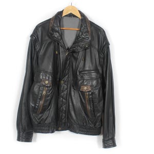 Könnte beinhalten: Schwarze Lederjacke mit braunem Kragen und Akzenten, hängt an einem Holzbügel. Die Jacke hat mehrere Taschen mit Reißverschlüssen und einen gerippten Saum. Klassischer Stil.