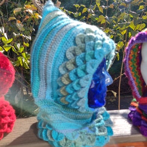 May include: A blue and green crocheted hooded cowl with a textured, scale-like pattern. The cowl is displayed on a wooden surface.