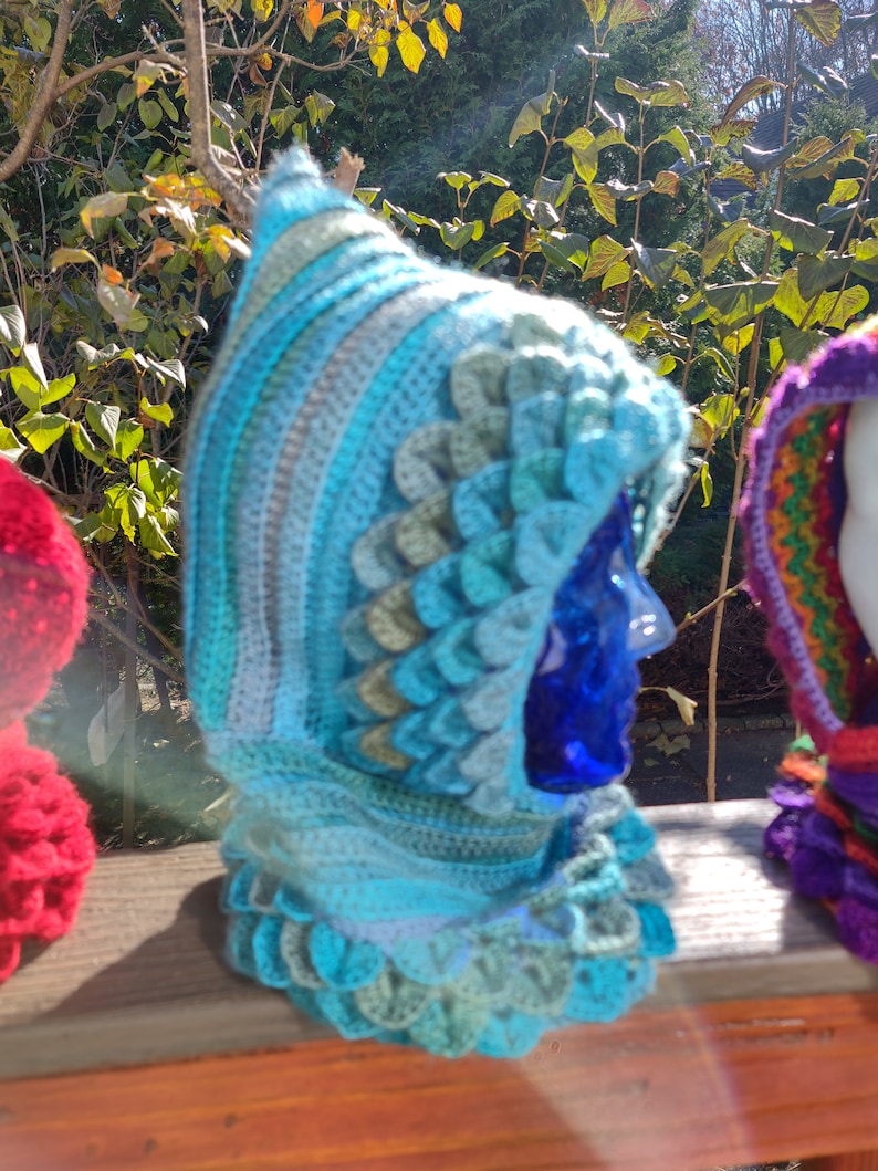 May include: A blue and green crocheted hooded cowl with a textured, scale-like pattern. The cowl is displayed on a wooden surface.