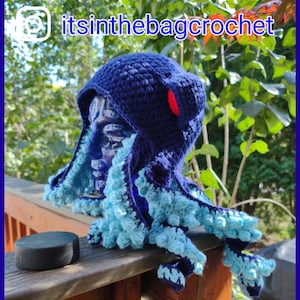 May include: A blue and turquoise crocheted octopus hat with a red pom-pom on top. The hat is worn on a mannequin head and the tentacles are draped over a wooden railing. The text "itsinthebagcrochet" is visible in the top right corner of the image.