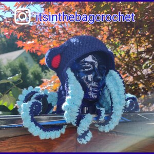 May include: A blue crocheted octopus hat with a red pom-pom on top. The hat is worn on a mannequin head and has blue and white tentacles. The hat is sitting on a black and silver surface. The text "itsinthebagcrochet" is visible in the image.