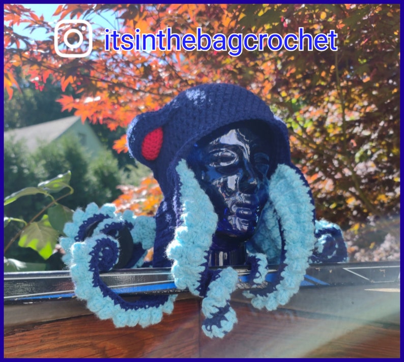 May include: A blue crocheted octopus hat with a red pom-pom on top. The hat is worn on a mannequin head and has blue and white tentacles. The hat is sitting on a black and silver surface. The text "itsinthebagcrochet" is visible in the image.