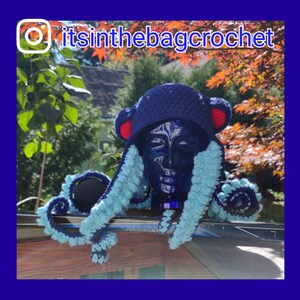 May include: A blue crocheted octopus hat with red ears and blue and light blue tentacles. The hat is on a wooden surface with a green leafy background.