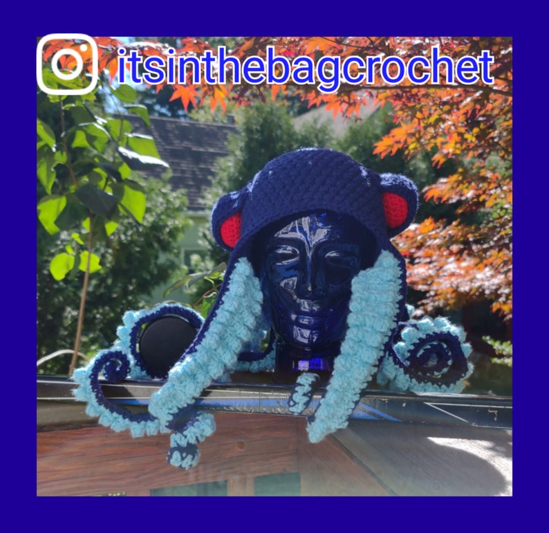 May include: A blue crocheted octopus hat with red ears and blue and light blue tentacles. The hat is on a wooden surface with a green leafy background.