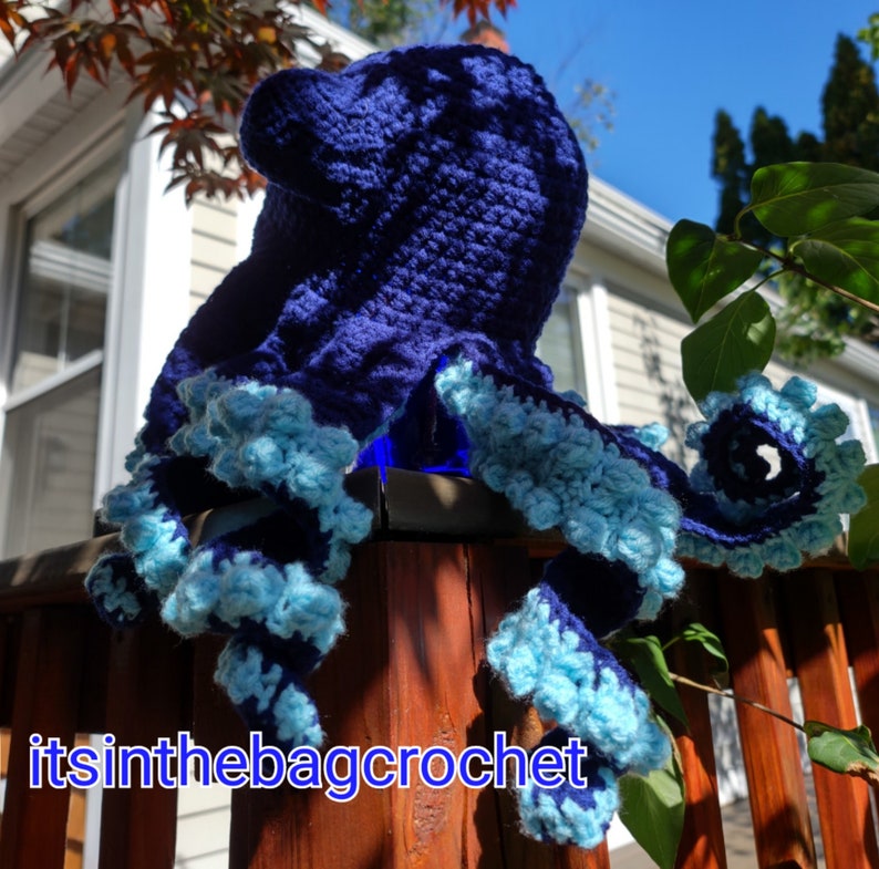 May include: A crocheted octopus with blue and light blue yarn. The octopus is sitting on a wooden fence.