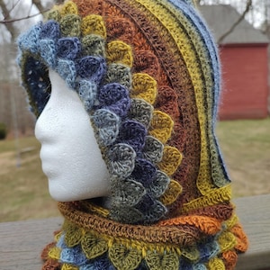 May include: A crocheted hooded cowl with a pointed top. The cowl is made in a variety of shades of brown, yellow, green, and blue. The cowl has a textured, scaly pattern.