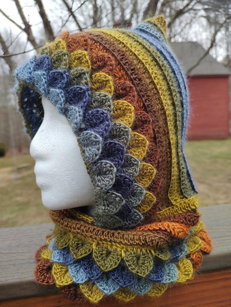 May include: A crocheted hooded cowl with a pointed top. The cowl is made in a variety of shades of brown, yellow, green, and blue. The cowl has a textured, scaly pattern.