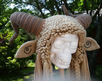 Puede incluir: Un gorro de crochet con cuernos y orejas, que se asemeja a una criatura mítica. El gorro es beige y marrón con una superficie texturizada. El gorro se lleva sobre un cráneo blanco.